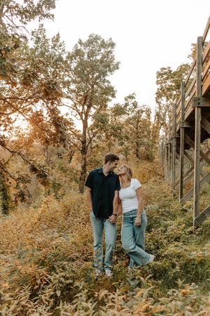 A couple stands together in an autumn forest setting with golden foliage and wooden fence posts in the background.