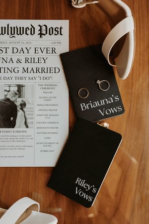 Wedding details including vow books and newspaper-style invitation laid on wooden surface with white ribbons.