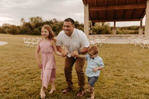 A family walks together across a sunlit grassy field with white chairs visible in the background.