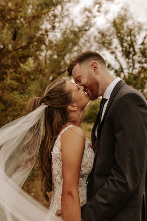 A couple in wedding attire share an intimate moment in a wooded outdoor setting as a veil flows in the breeze.