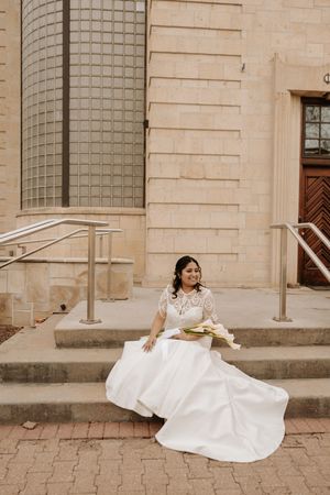 A bride in a flowing white dress sits gracefully on stone steps outside a historic building with columns.