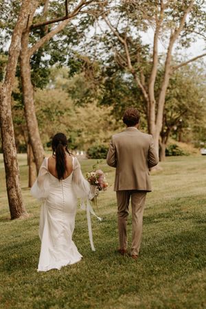 A couple walks through a serene wooded area on their wedding day, with the bride in white and groom in tan.