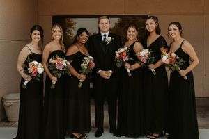 Wedding party members stand together in elegant black attire against a neutral brown wall backdrop.