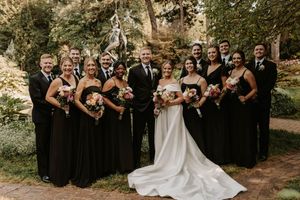 Wedding party in formal black attire and white dress pose together in an outdoor garden setting at sunset.
