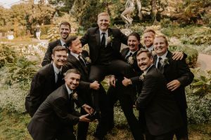 A group of groomsmen in black suits pose playfully together outdoors in a natural garden setting.