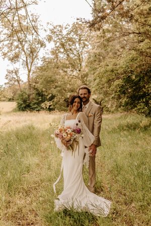 A romantic wedding couple embraces in a sun-drenched meadow with wildflowers and trees creating a dreamy backdrop.