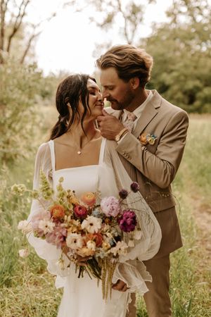 Wedding couple poses in a field with colorful bouquet of pink and burgundy flowers.