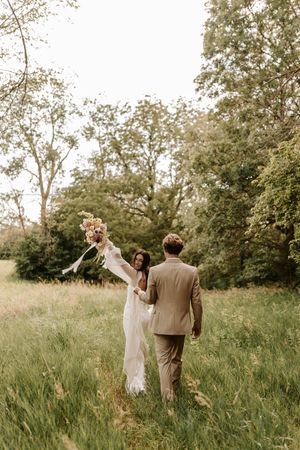 A couple celebrates their wedding day in a sunlit meadow surrounded by trees while holding hands.