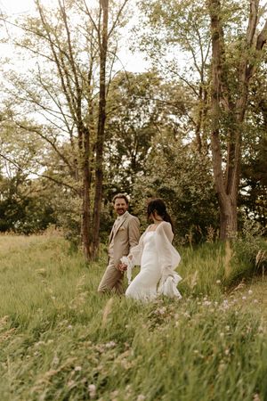Newlyweds stroll together through tall grass and trees in a romantic outdoor setting.