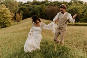 A couple in wedding attire runs playfully through a sunlit grassy hillside at golden hour.