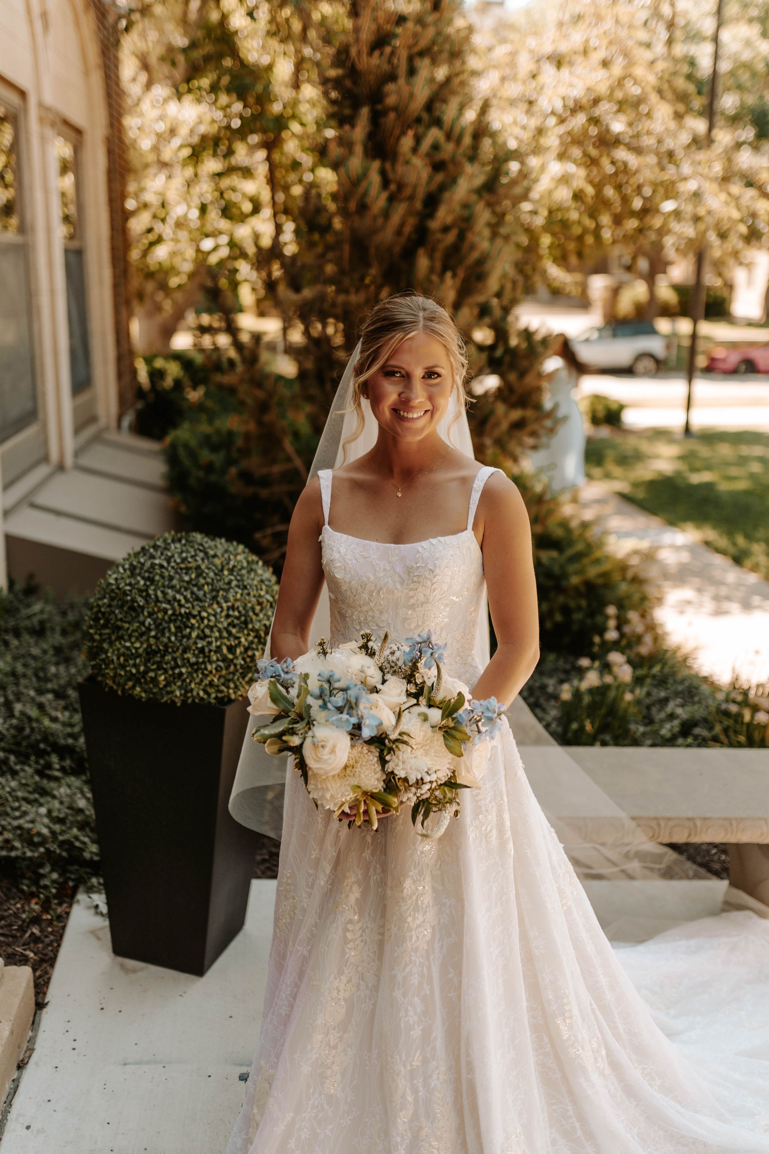 Bride in a flowing white dress holding a bouquet stands in front of landscaped garden and building.