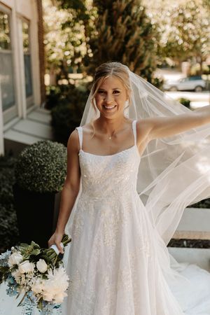 Joyful bride in lace gown twirls with flowing veil outdoors on sunny wedding day.