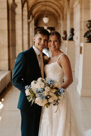 Elegant wedding couple shares intimate moments in a columned hallway with white and blue floral bouquet.