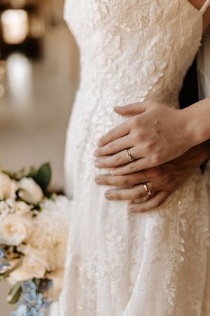 Close-up shots of hands wearing wedding rings against a white lace dress with delicate floral bouquet details.