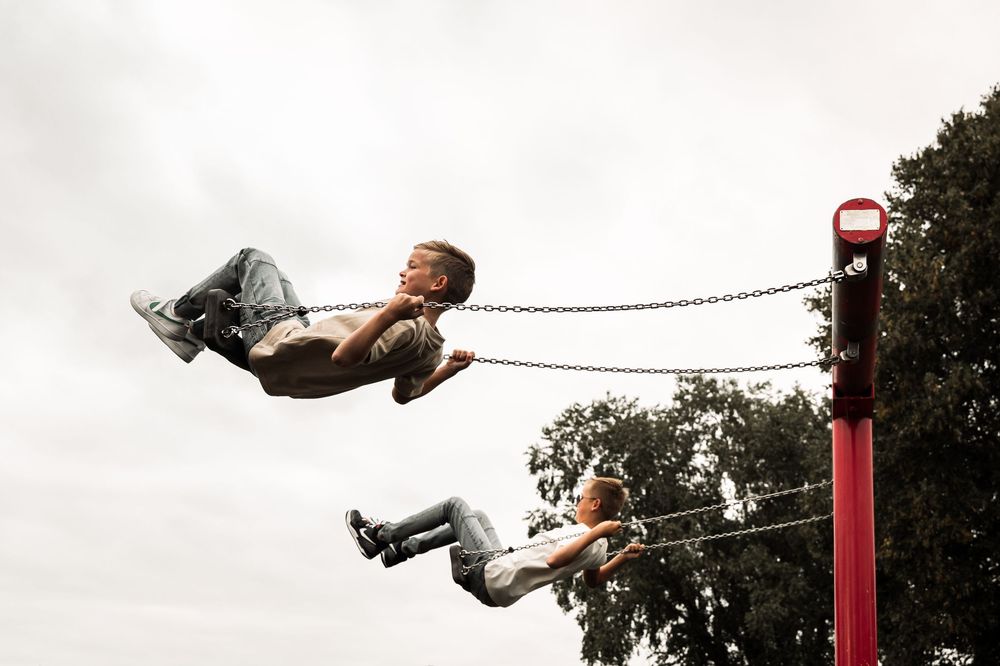 A child swings high in the air on a playground swing set against a cloudy sky backdrop.