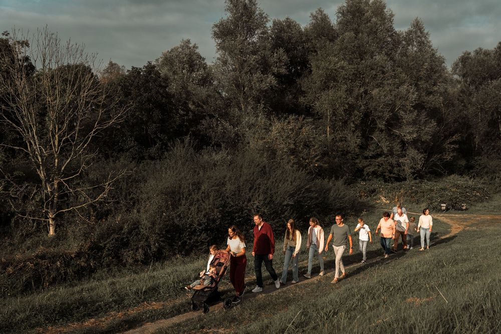 A group of people walk in a line along a grassy hillside path at dusk with trees in the background.