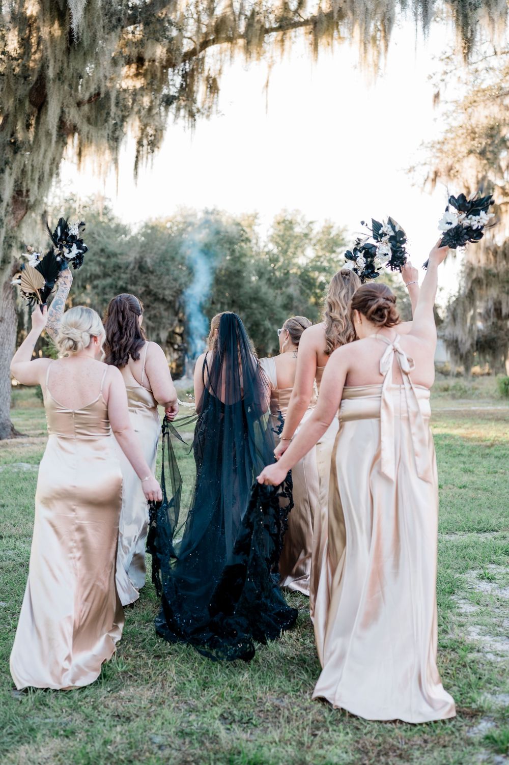 Bridesmaids in champagne dresses and one in black pose together on grassy lawn with Spanish moss hanging from trees.