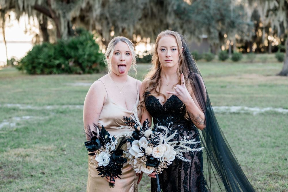 A group of bridesmaids in black dresses pose together outdoors with black and white pom poms.