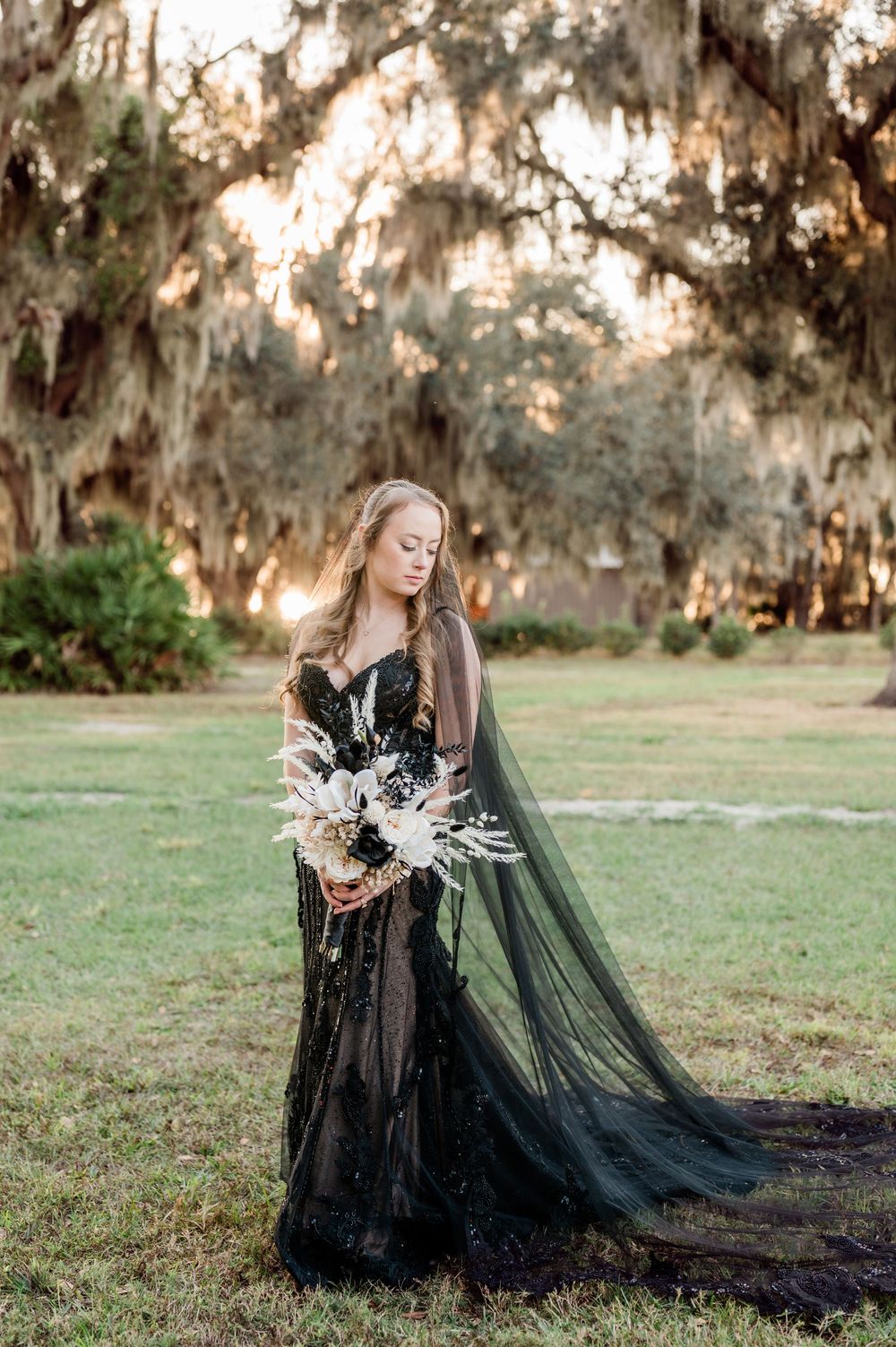 Artistic photoshoot in a field with Spanish moss trees featuring a black gothic wedding dress and white floral bouquet.