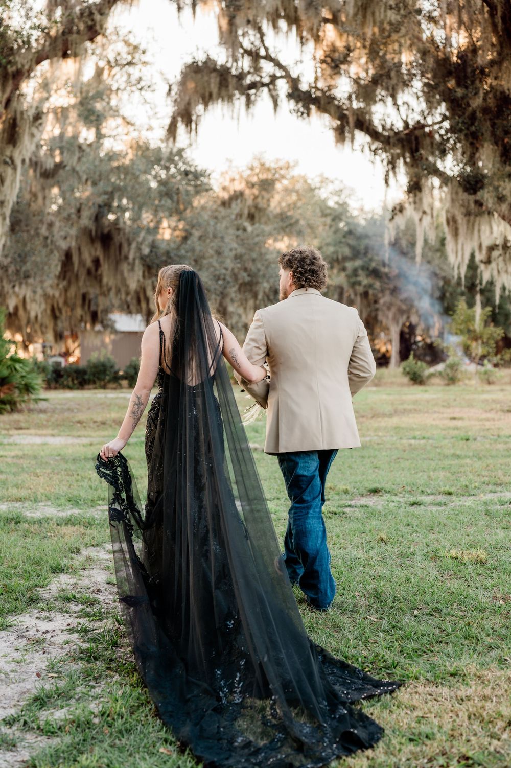 A couple walks through a scenic garden with Spanish moss as the bride's dramatic black veil trails behind them.