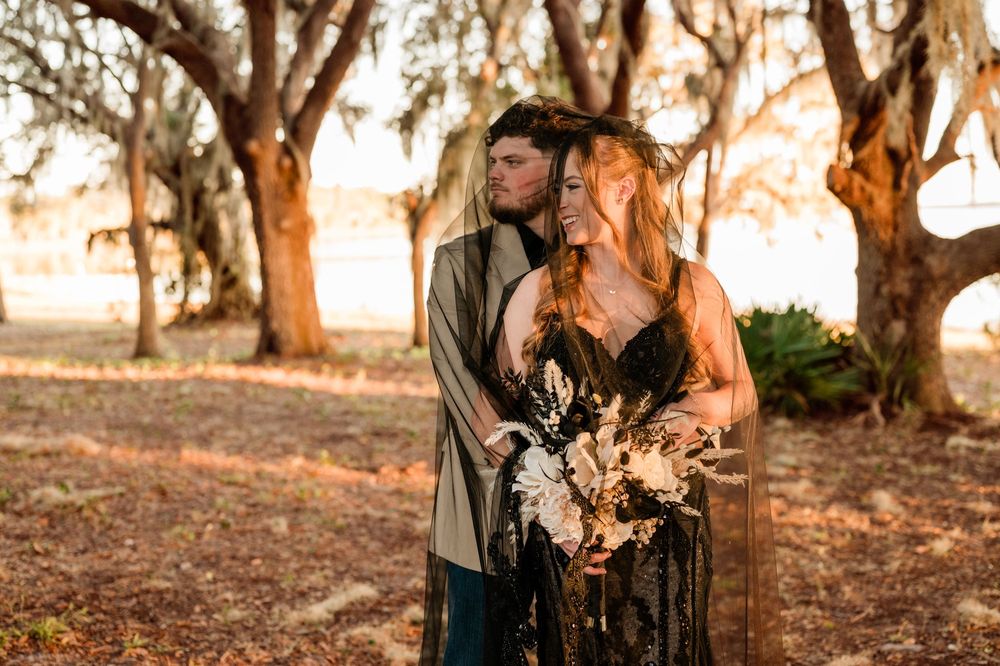A couple stands together in a romantic sunset setting among large trees while holding a dried floral bouquet.