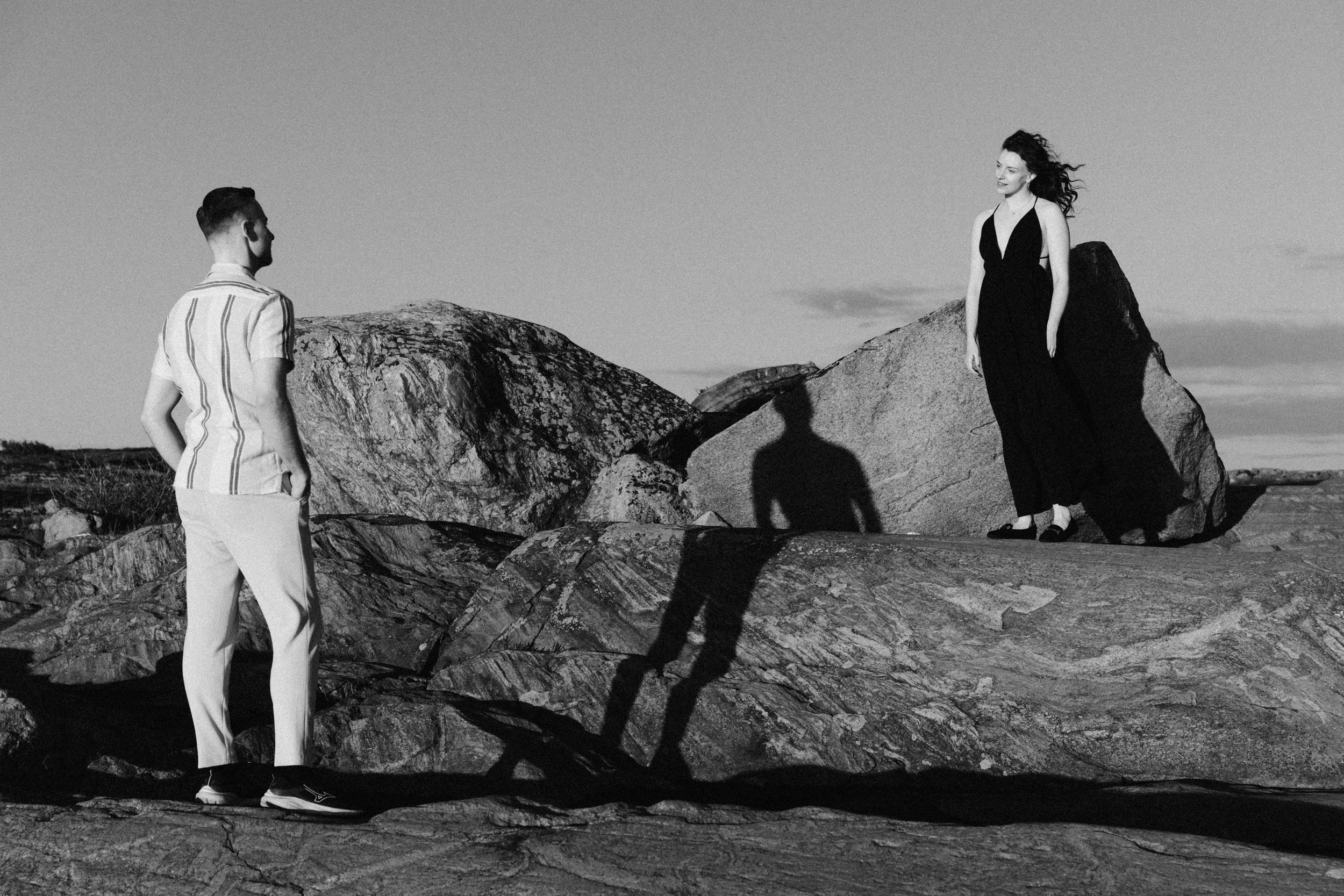 Two figures in contrasting black and white outfits stand on rocky terrain against a dramatic sky.