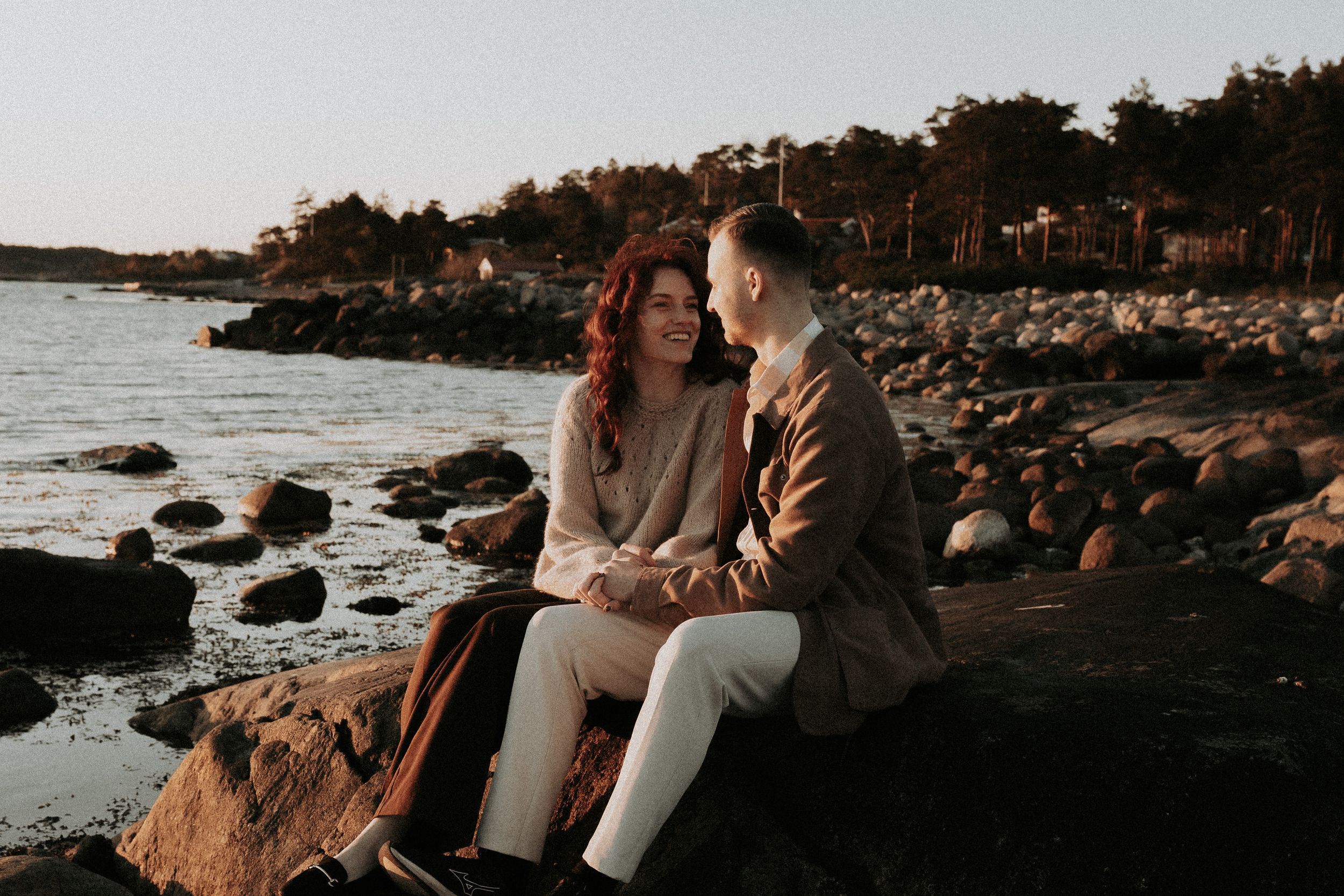 Couple sitting together on coastal rocks at sunset with pine trees in background.