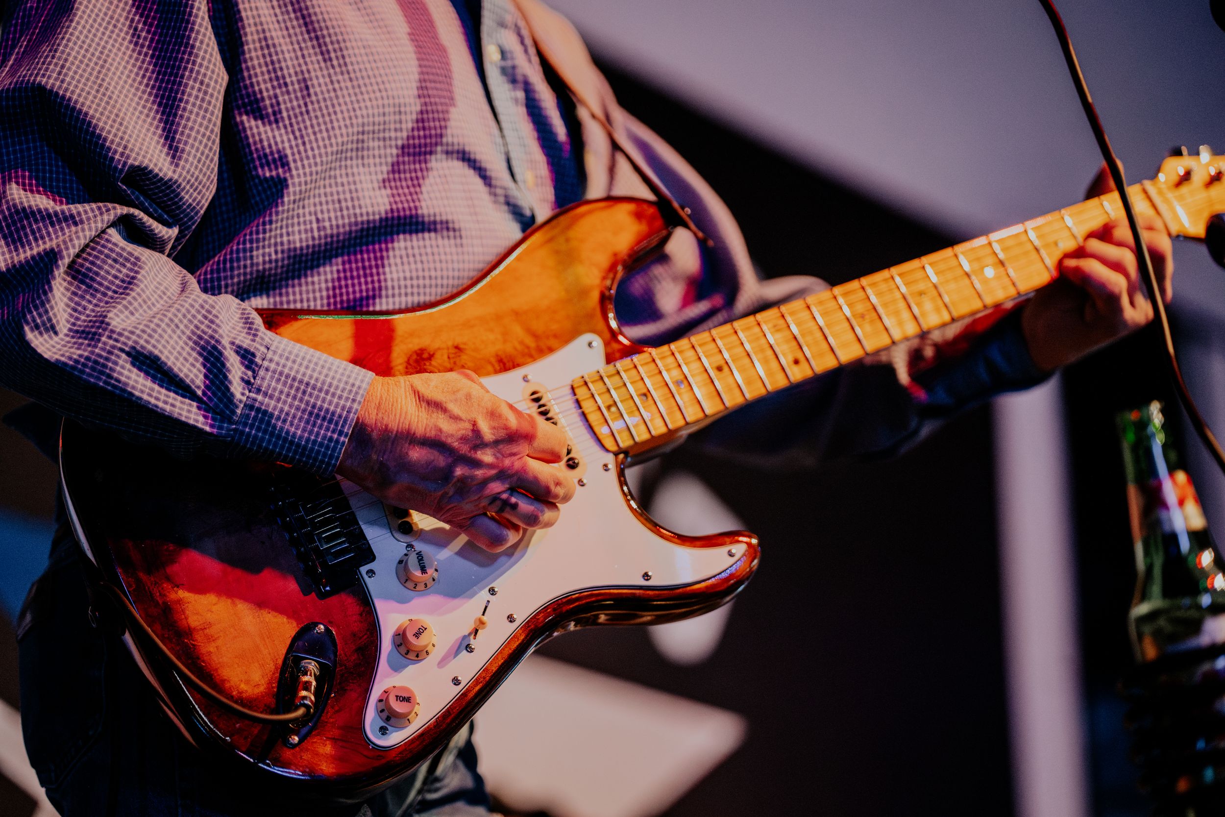 Close-up shot of hands playing an orange and white electric guitar under purple stage lighting.