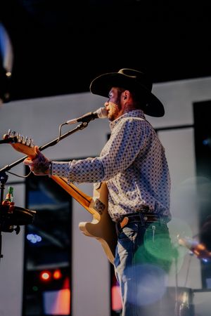 Country musician in cowboy hat and patterned shirt sings while playing guitar on stage.
