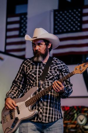 Bass guitarist in plaid shirt and cowboy hat performs in front of American flag backdrop.