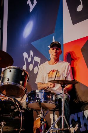 Drummer performs behind drum kit with Texas-themed stage backdrop featuring musical notes.