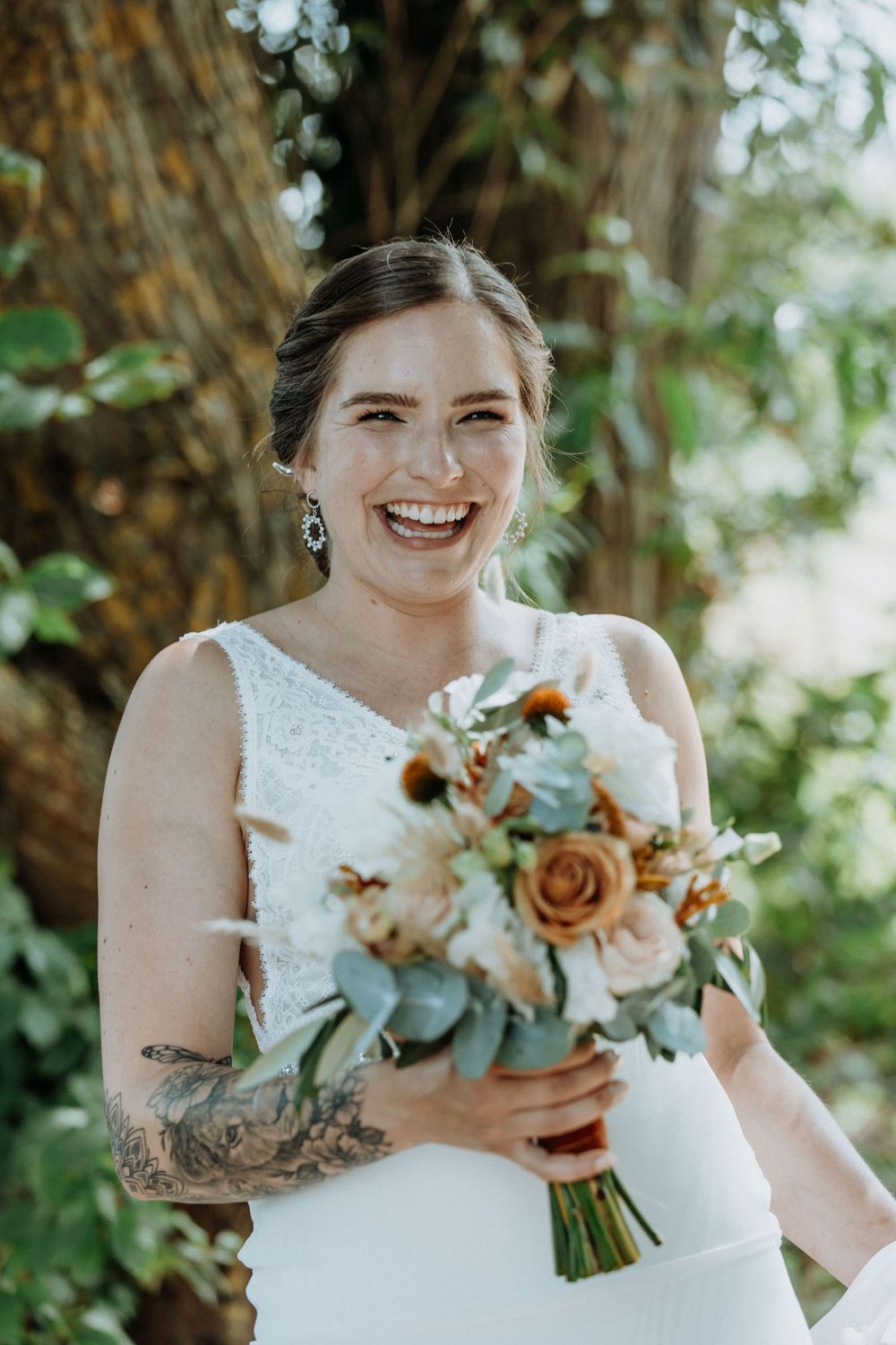 A joyful bride in a white dress holds a beautiful bouquet with orange roses and eucalyptus against a natural backdrop.