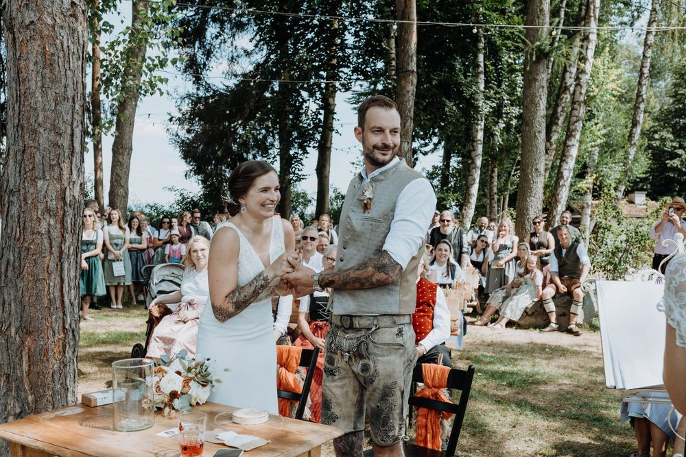 Outdoor wedding ceremony setup with guests gathered among tall trees in a natural woodland setting.