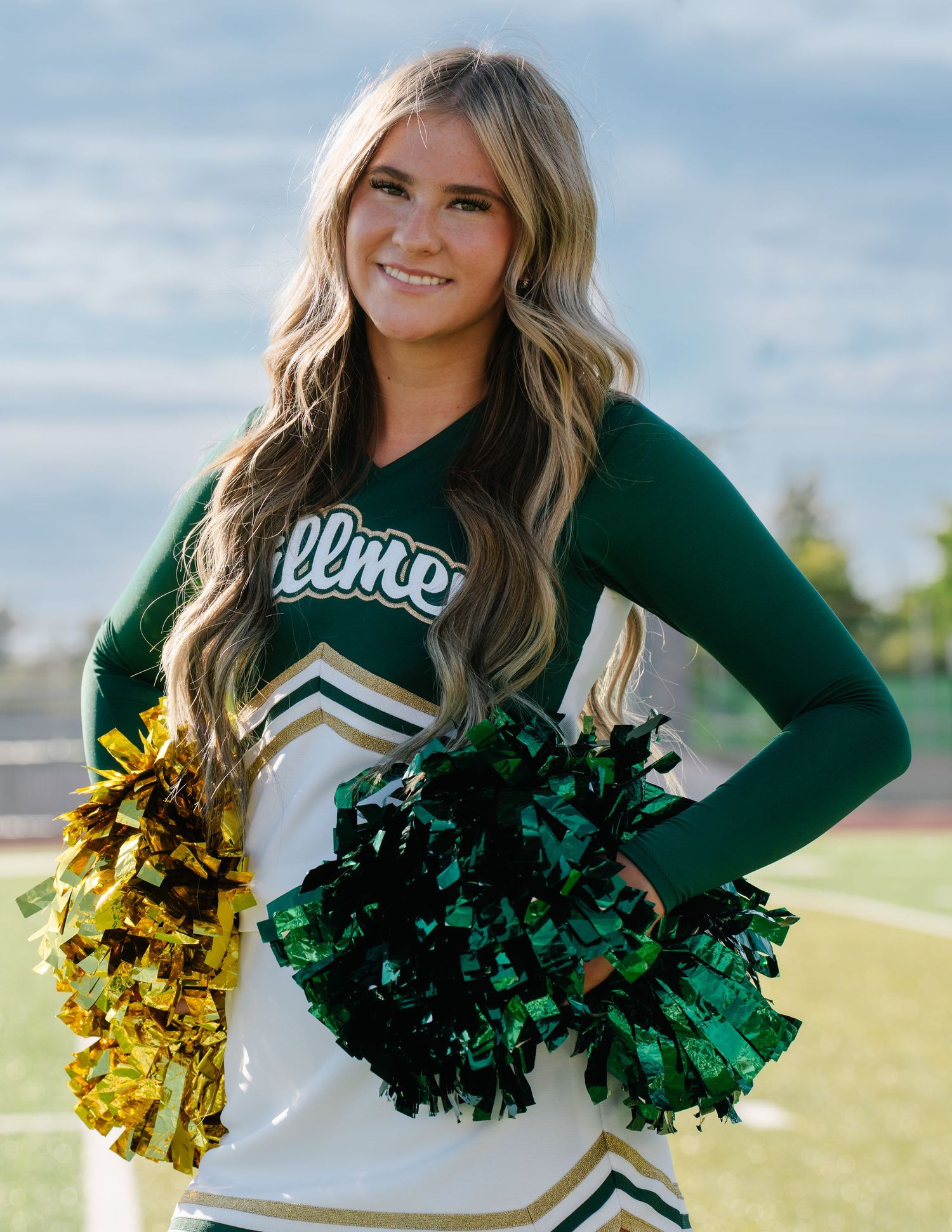 Smiling cheerleader in green and white uniform holding gold and green pom-poms on a football field.