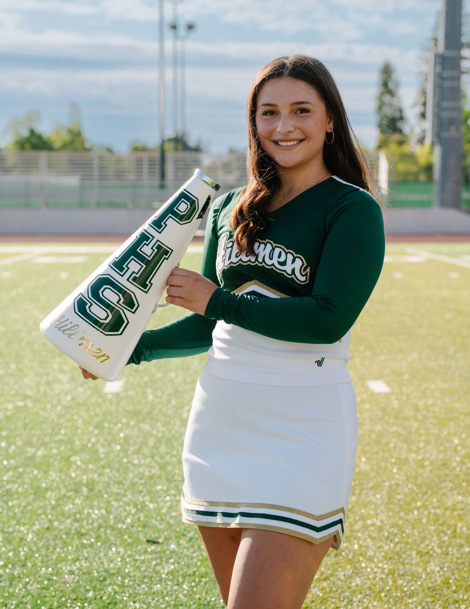 Cheerleader in green and white uniform holding a PHS megaphone on a football field.