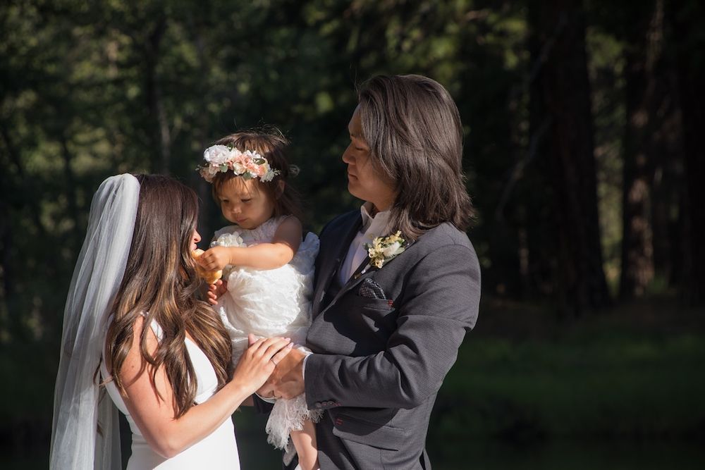 A couple in wedding attire stands together in a wooded setting holding a small child wearing a floral crown.