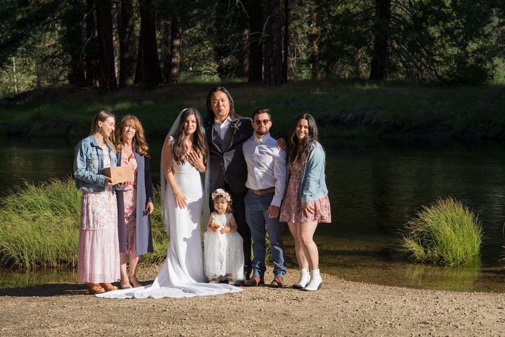 A group wearing white and light colored clothing poses outdoors in a natural setting near trees and water.