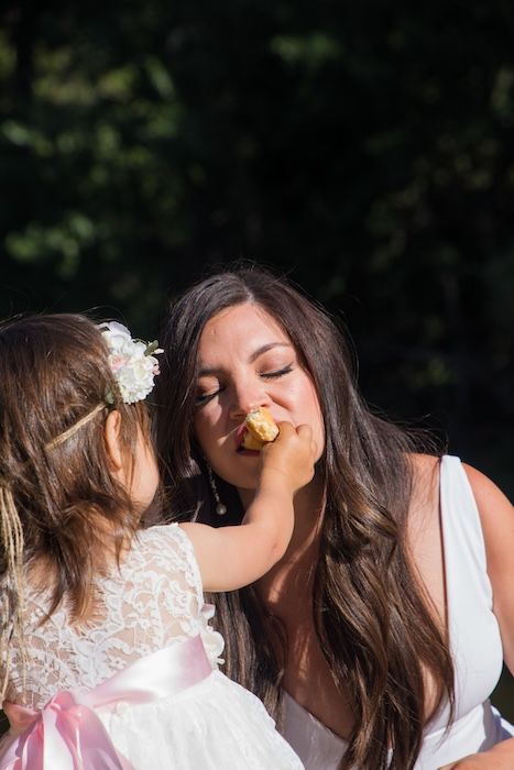 A young child in a white lace dress playfully touches someone's face in a sunny outdoor setting.