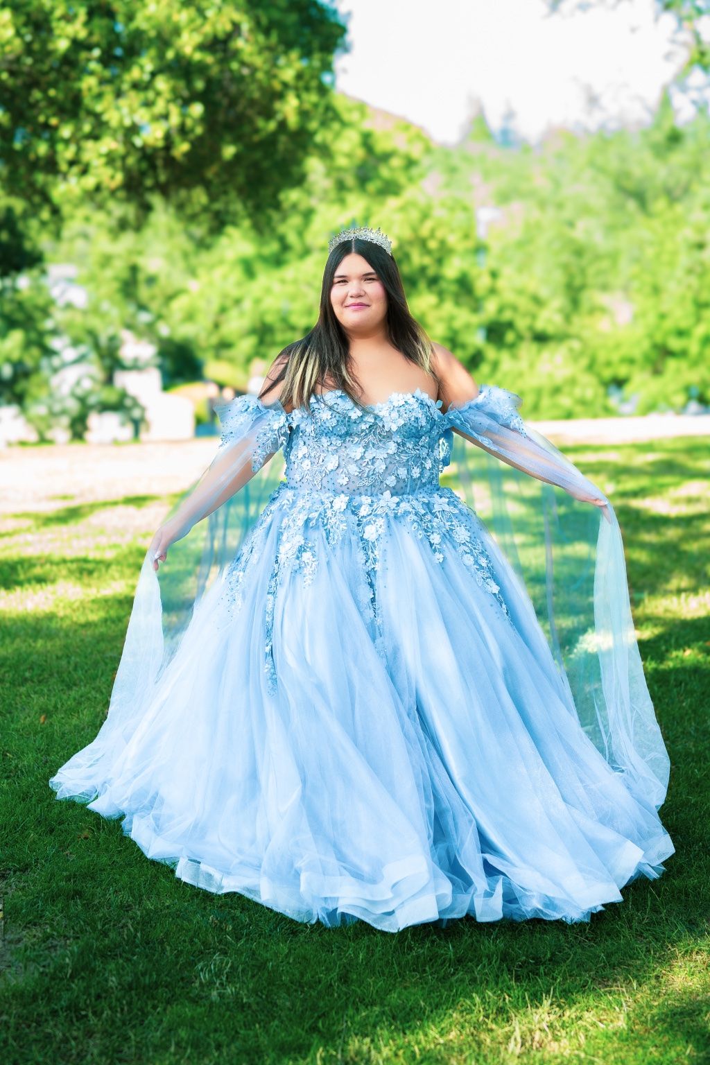 A flowing light blue ballgown with floral details twirls gracefully in a sunny green park setting.
