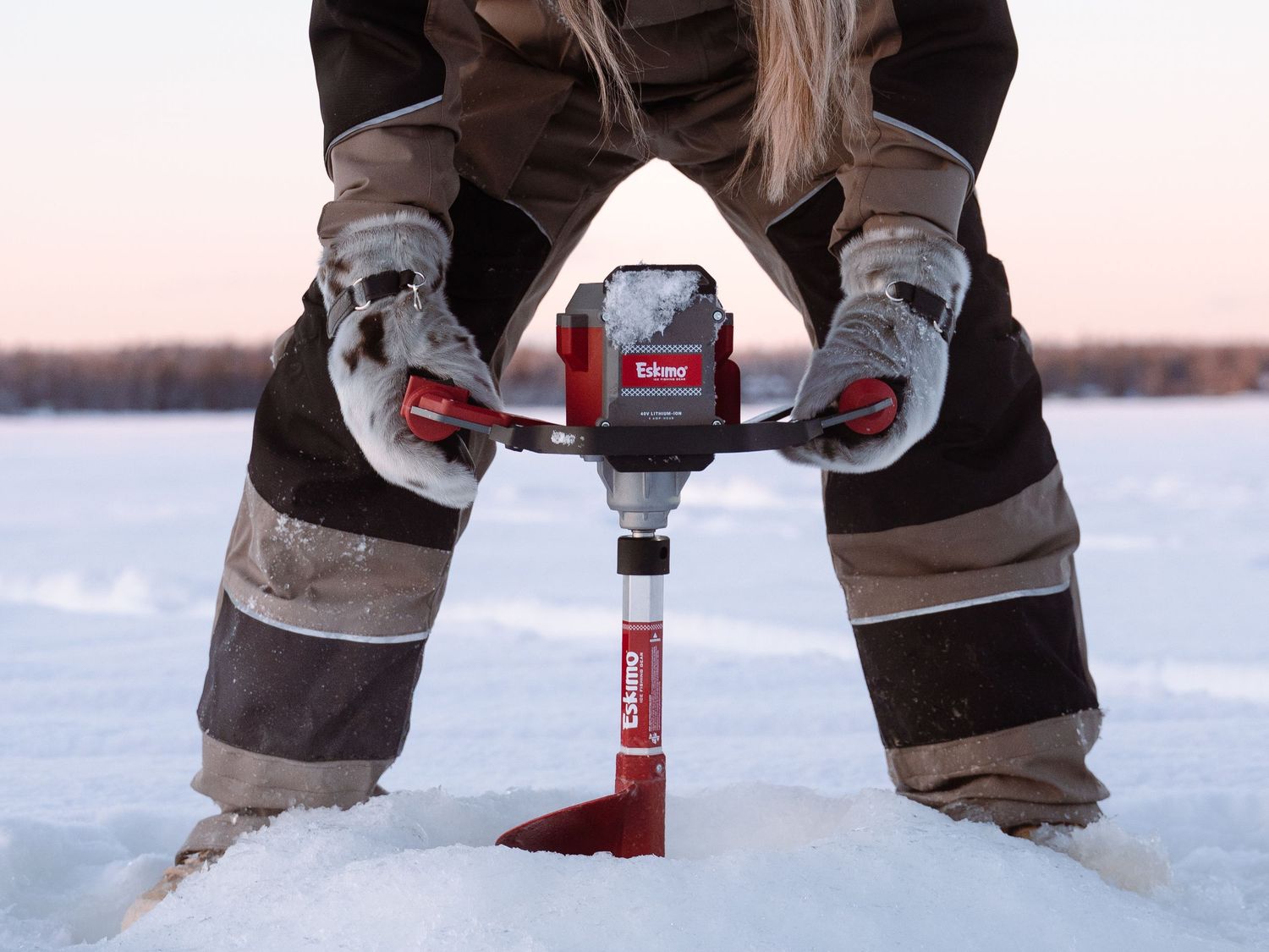Close-up view of hands holding red ice fishing auger in winter snow.
