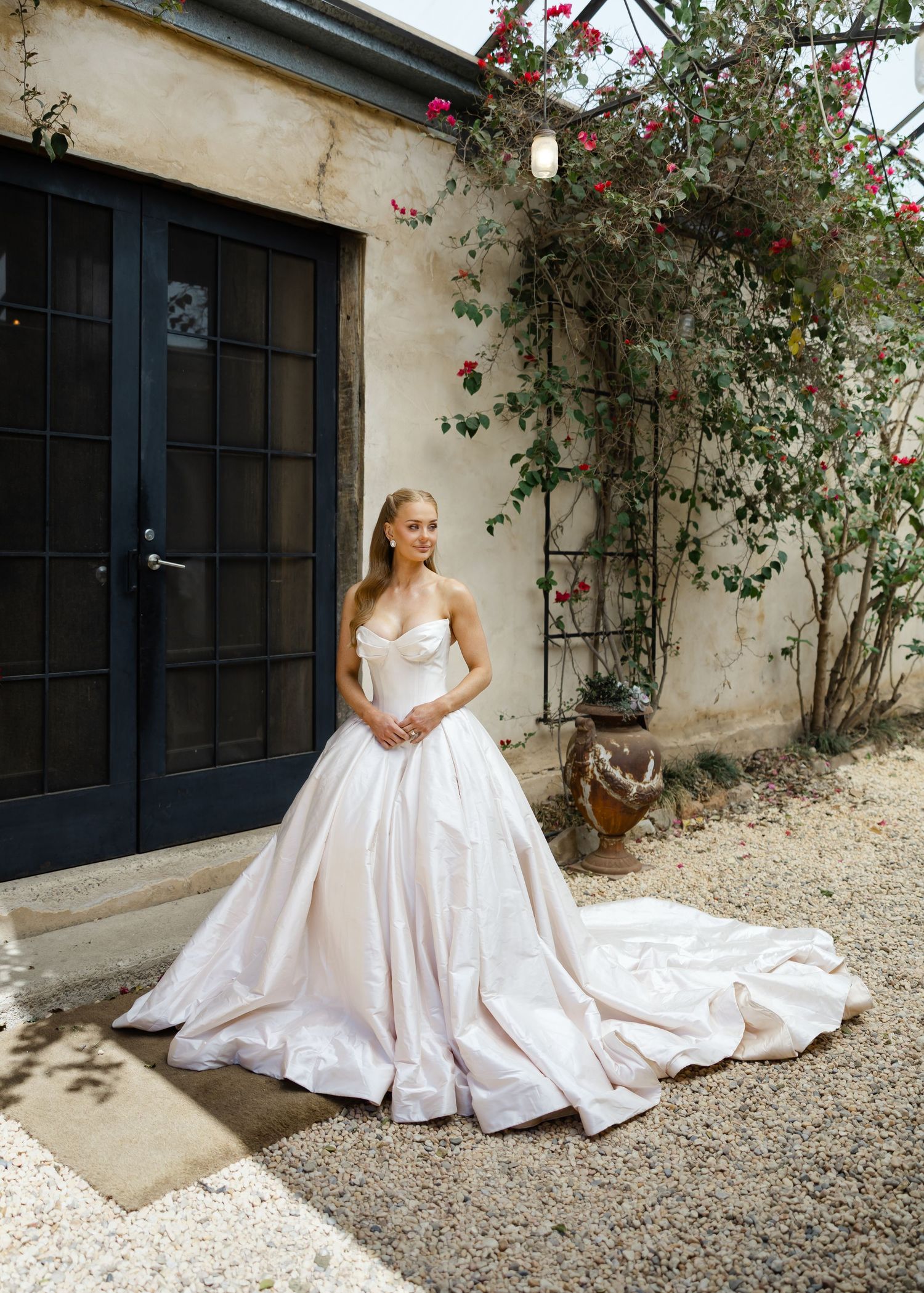 A white wedding gown with a flowing train spreads elegantly across a rustic stone courtyard with climbing roses at Al Ru Farm in Adelaide