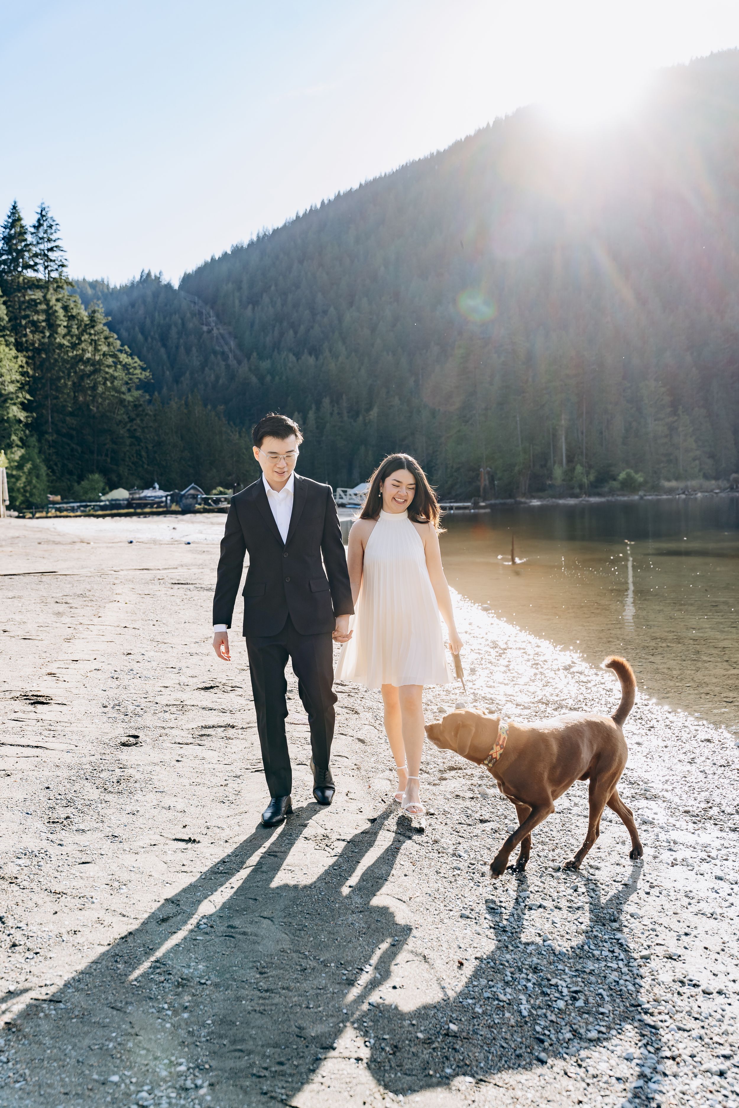 A couple walks along a scenic lakeside beach with their brown dog as sunlight streams through mountain trees.