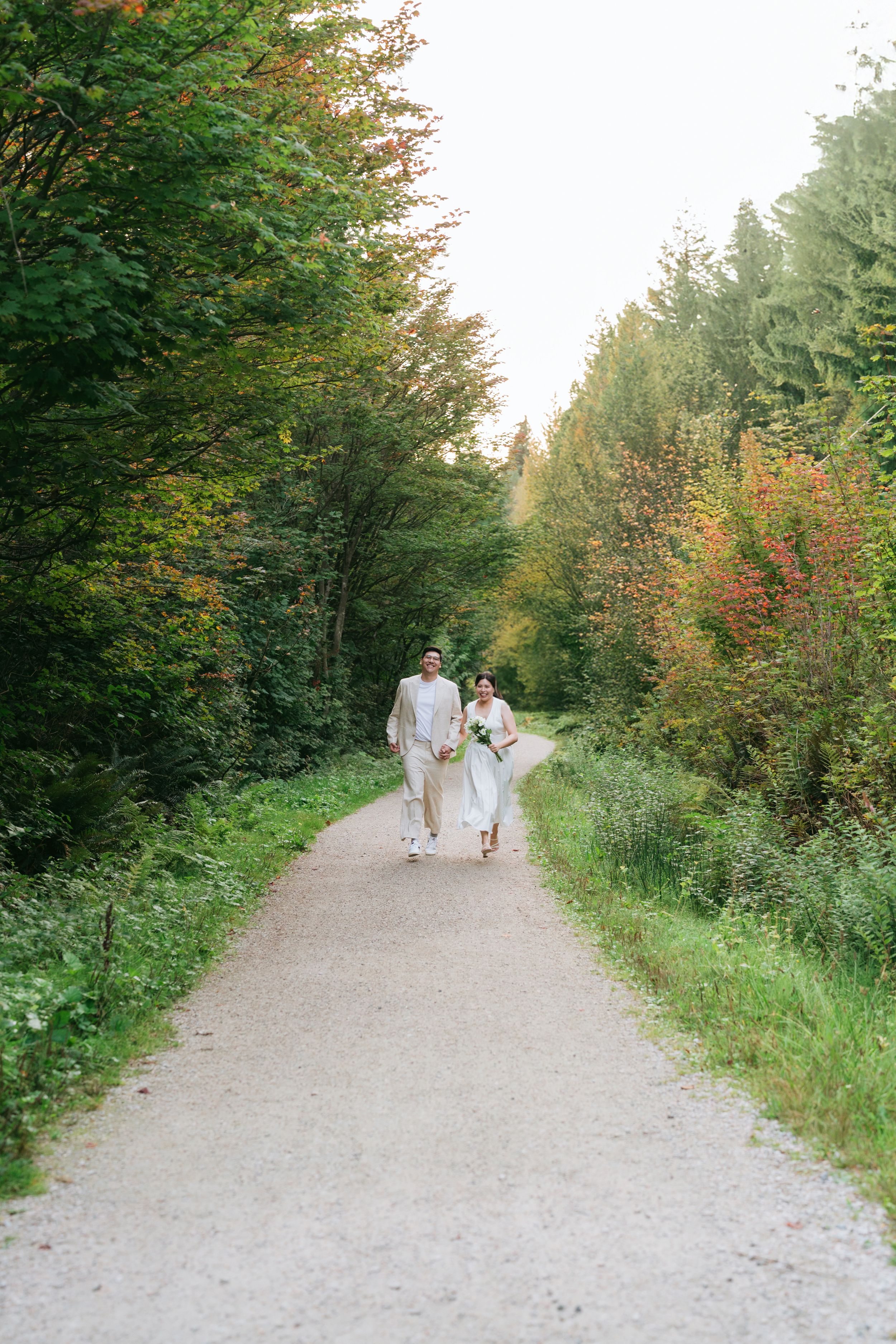 Two people in white dresses walk along a serene forest path lined with green trees and autumn foliage.