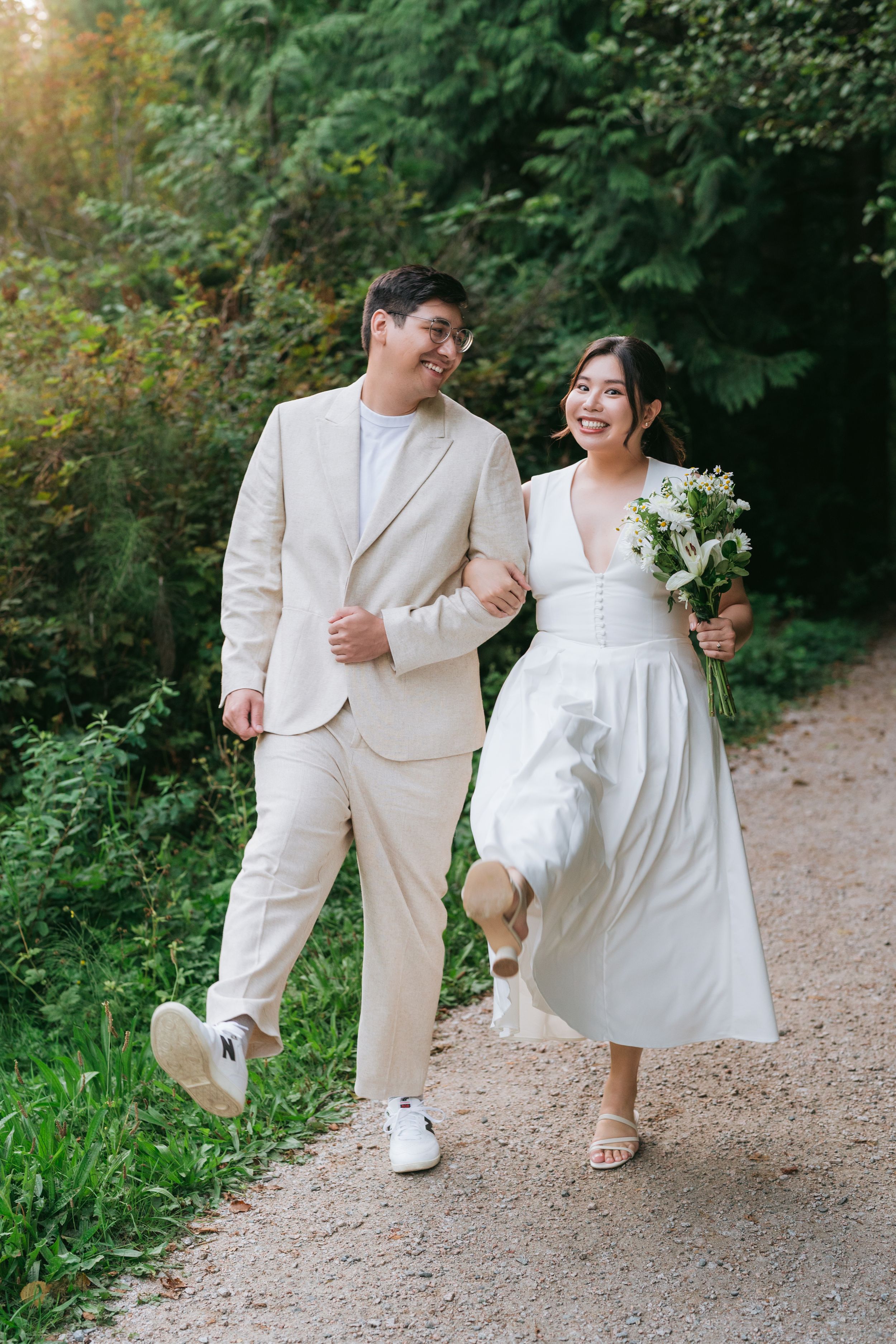 A couple in white attire walking and laughing together on a gravel path surrounded by lush greenery.