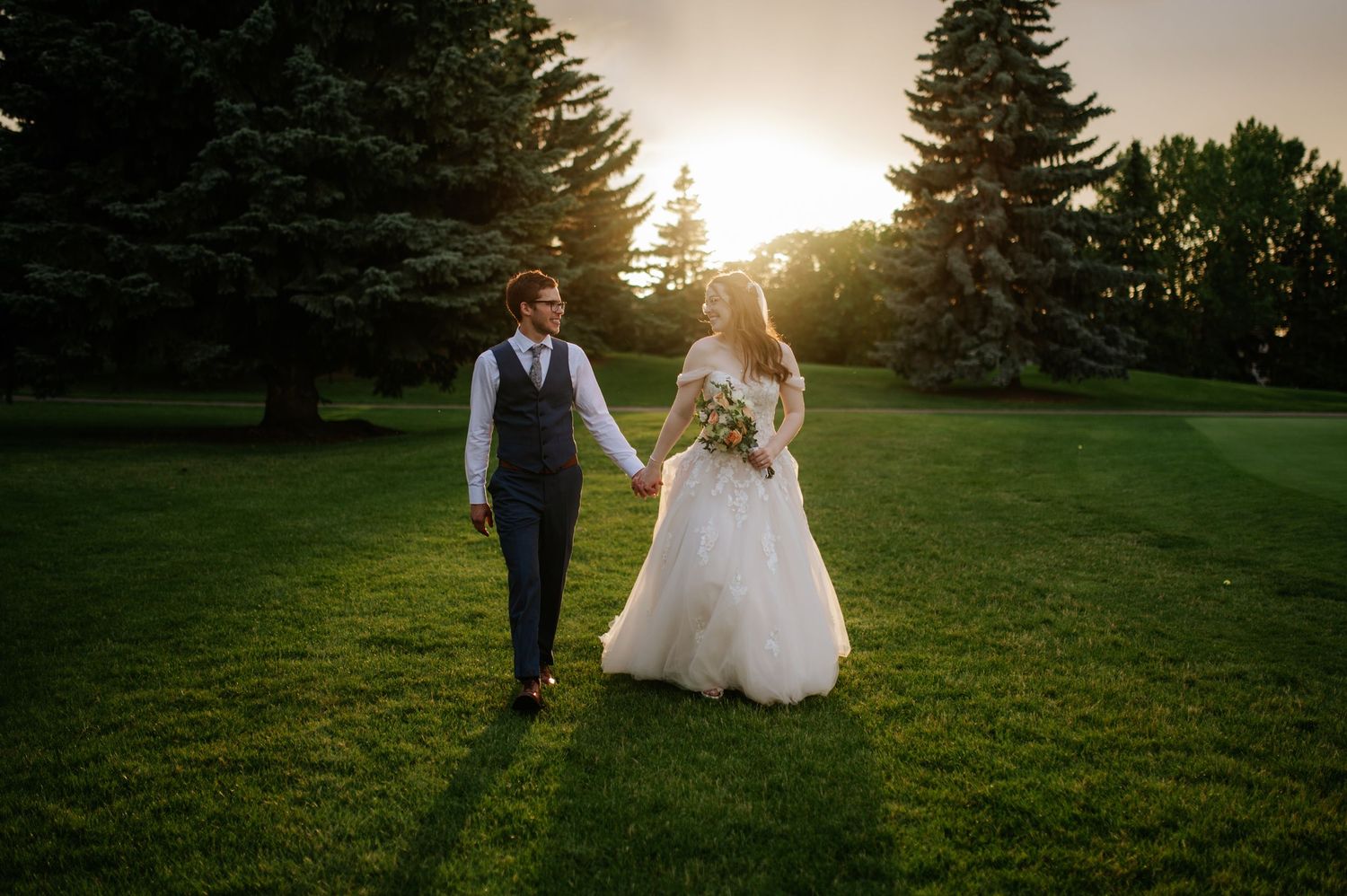 A couple walks hand in hand through a sunlit garden during golden hour as sunbeams filter through evergreen trees.