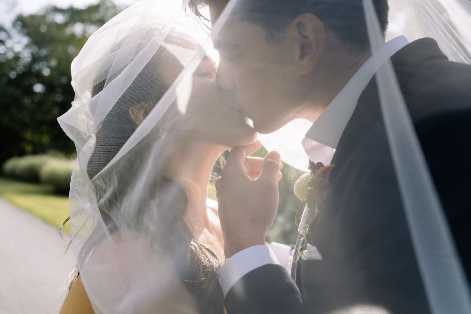 A romantic wedding moment captured through a delicate veil in soft sunlight outdoors.