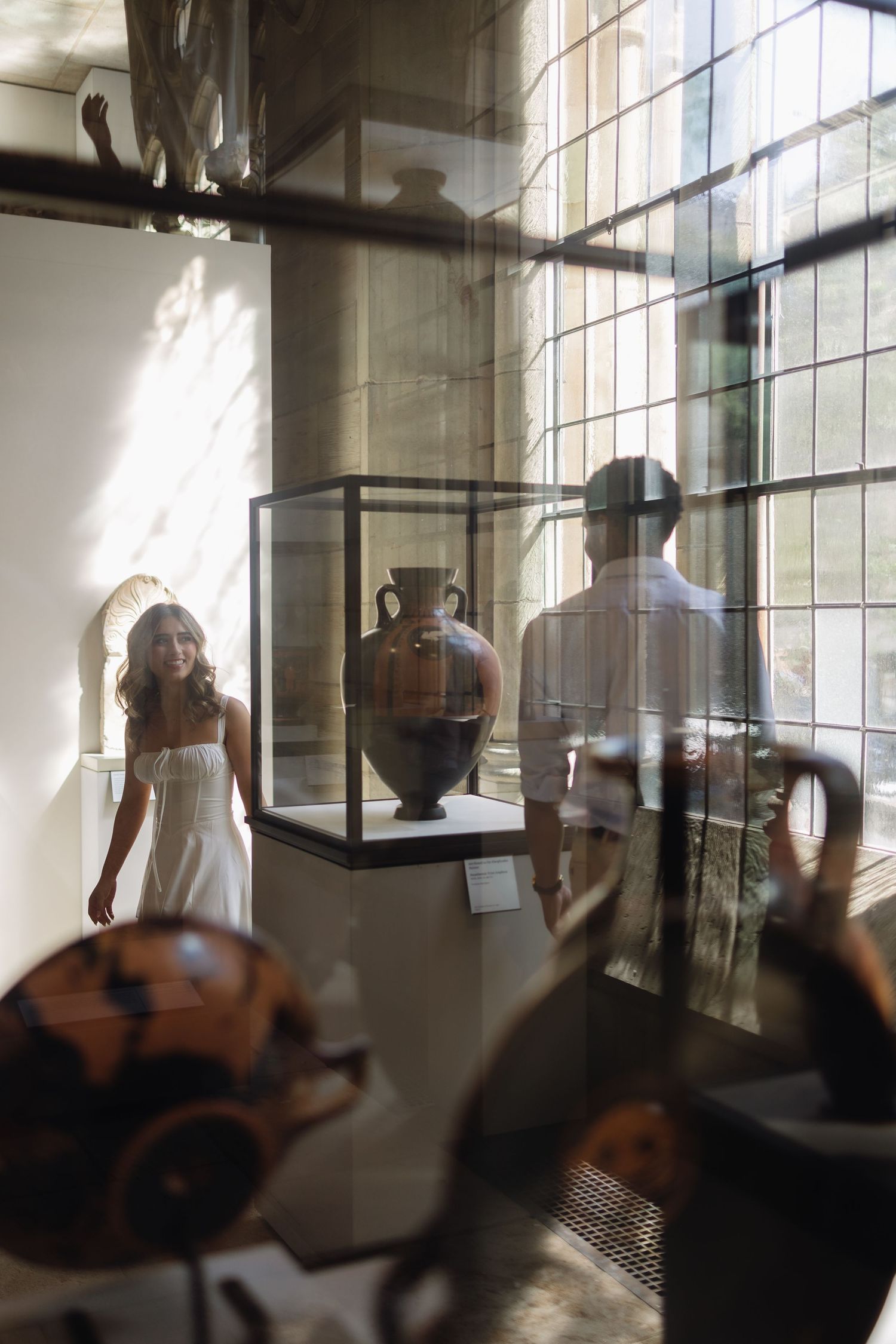 Silhouetted figures move through a sunlit gallery space with large windows and display cases containing ancient artifacts.