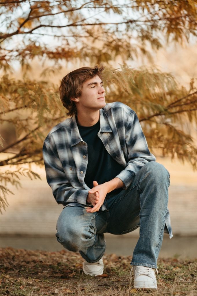 A person in a plaid flannel jacket and jeans crouches outdoors during autumn with golden foliage in the background.