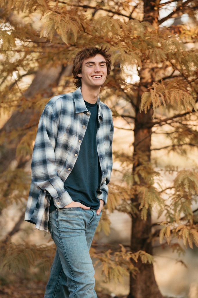 Person wearing plaid flannel shirt and jeans poses against autumn trees in golden light.