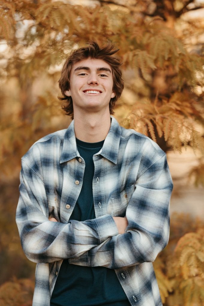 A person wearing a blue plaid flannel shirt poses against a backdrop of autumn foliage.